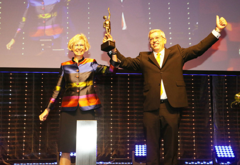 Anja and Roland Rüdinger stand on a stage and cheer to the audience. Roland holds up a golden trophy.