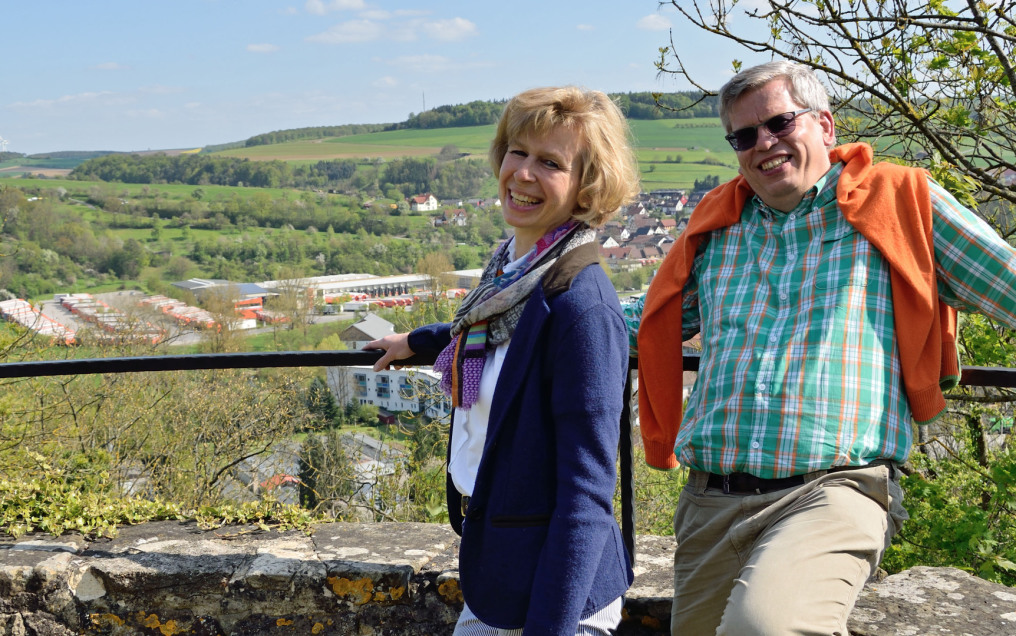 Anja and Roland Rüdinger laugh heartily to the viewer. They are standing on the tower of Krautheim Castle, of which only the railing is visible. In the background is the forwarding company grounds and all around a lot of nature: fields and forest.