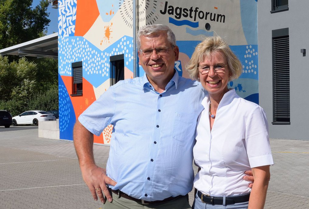 Anja and Roland Rüdinger laugh heartily to the viewer. They are standing on the tower of Krautheim Castle, of which only the railing is visible. In the background is the forwarding company grounds and all around a lot of nature: fields and forest.