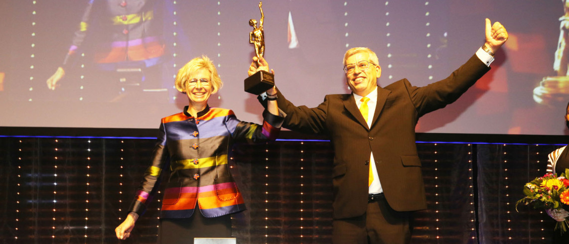 Anja and Roland Rüdinger hold up a golden trophy together. Both smile warmly. Roland also gives a "thumbs up".