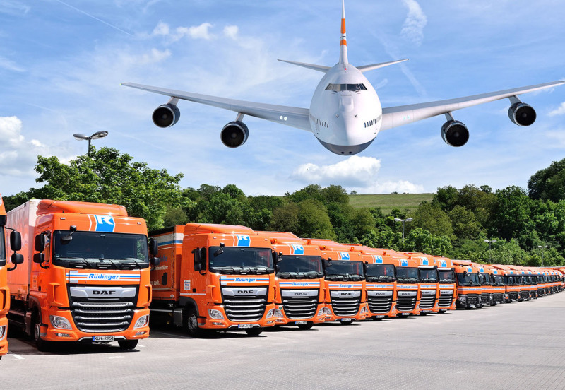Over a row of neatly parked tarpaulin low loaders on the left side, a cargo plane hovers in low-level flight towards the viewer. It is a photo collage.