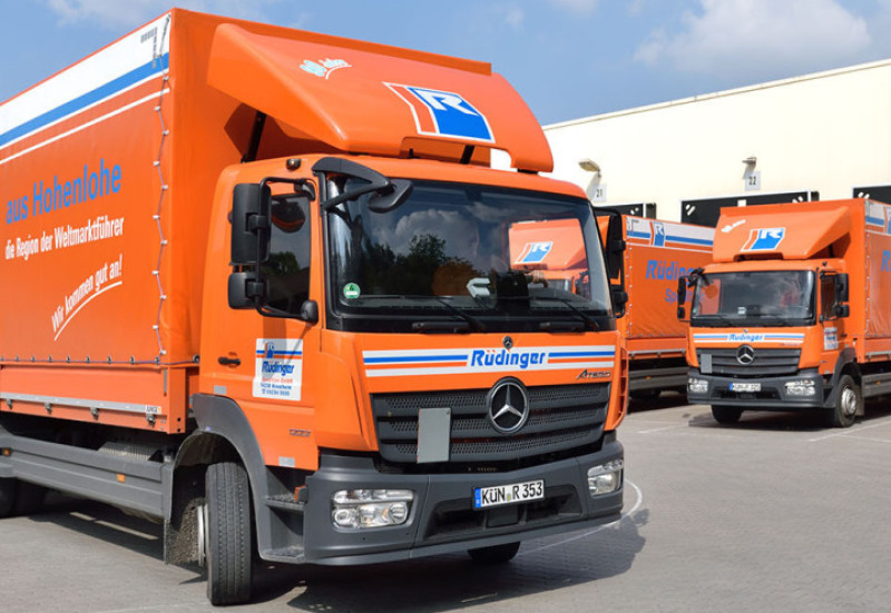 From the left, a 7.5-ton truck drives into the picture and towards the viewer, it is half visible. In the background, two more such trucks are parked at two ramps of the transshipment hall.